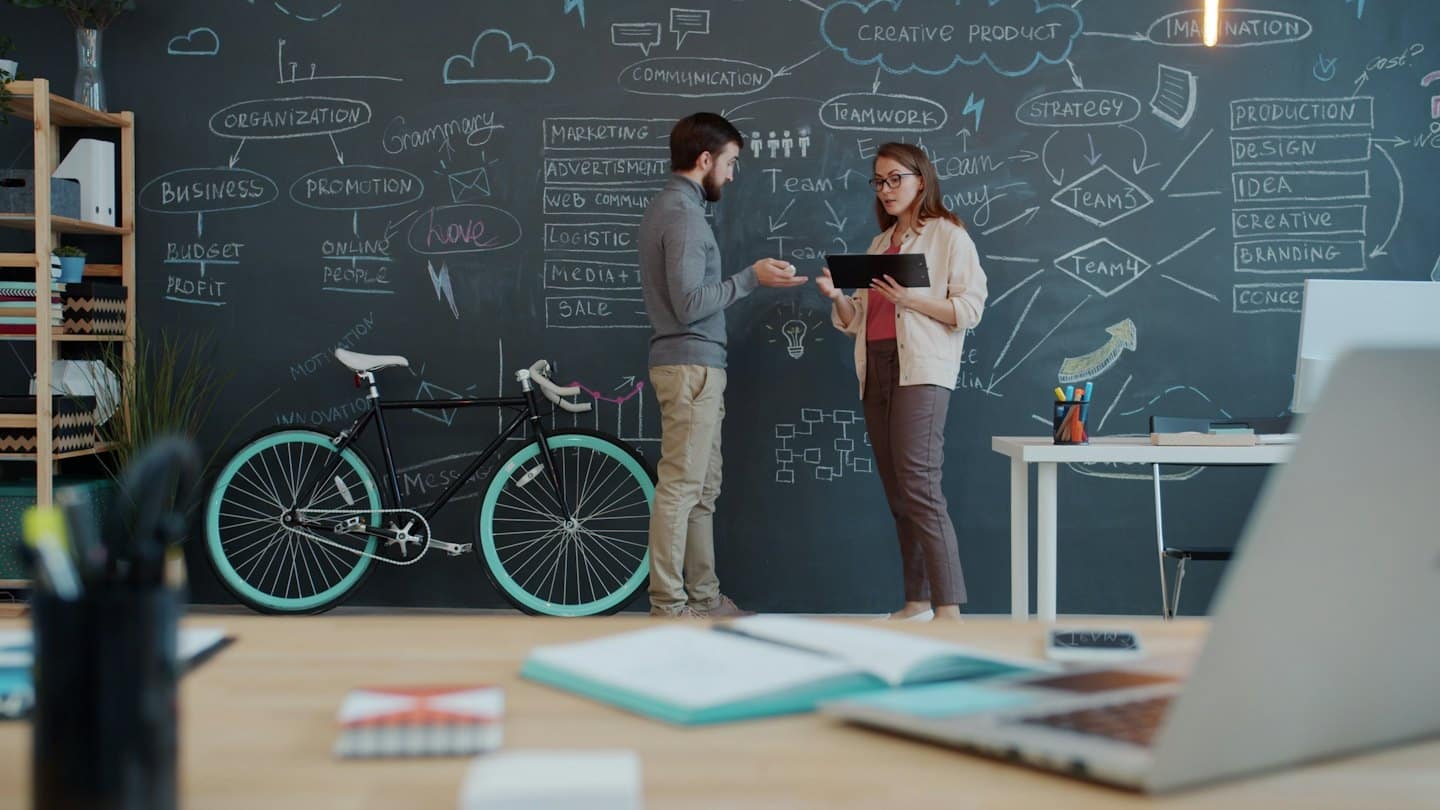 Twee professionals bespreken een businessplan met diagrammen op een schoolbord.